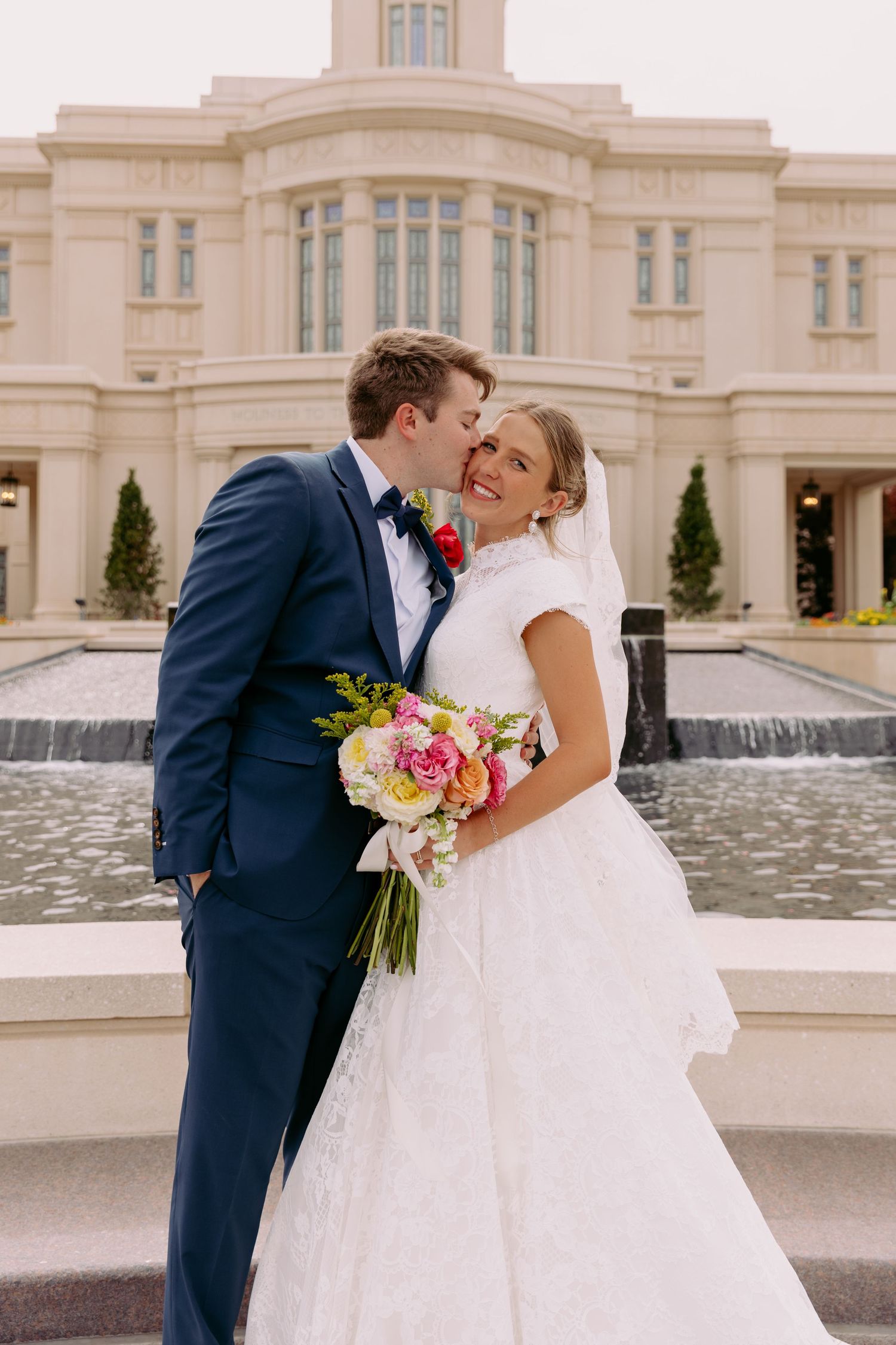 Bride and groom share a kiss outside a white temple, bride holding a colorful bouquet in a lace wedding gown.