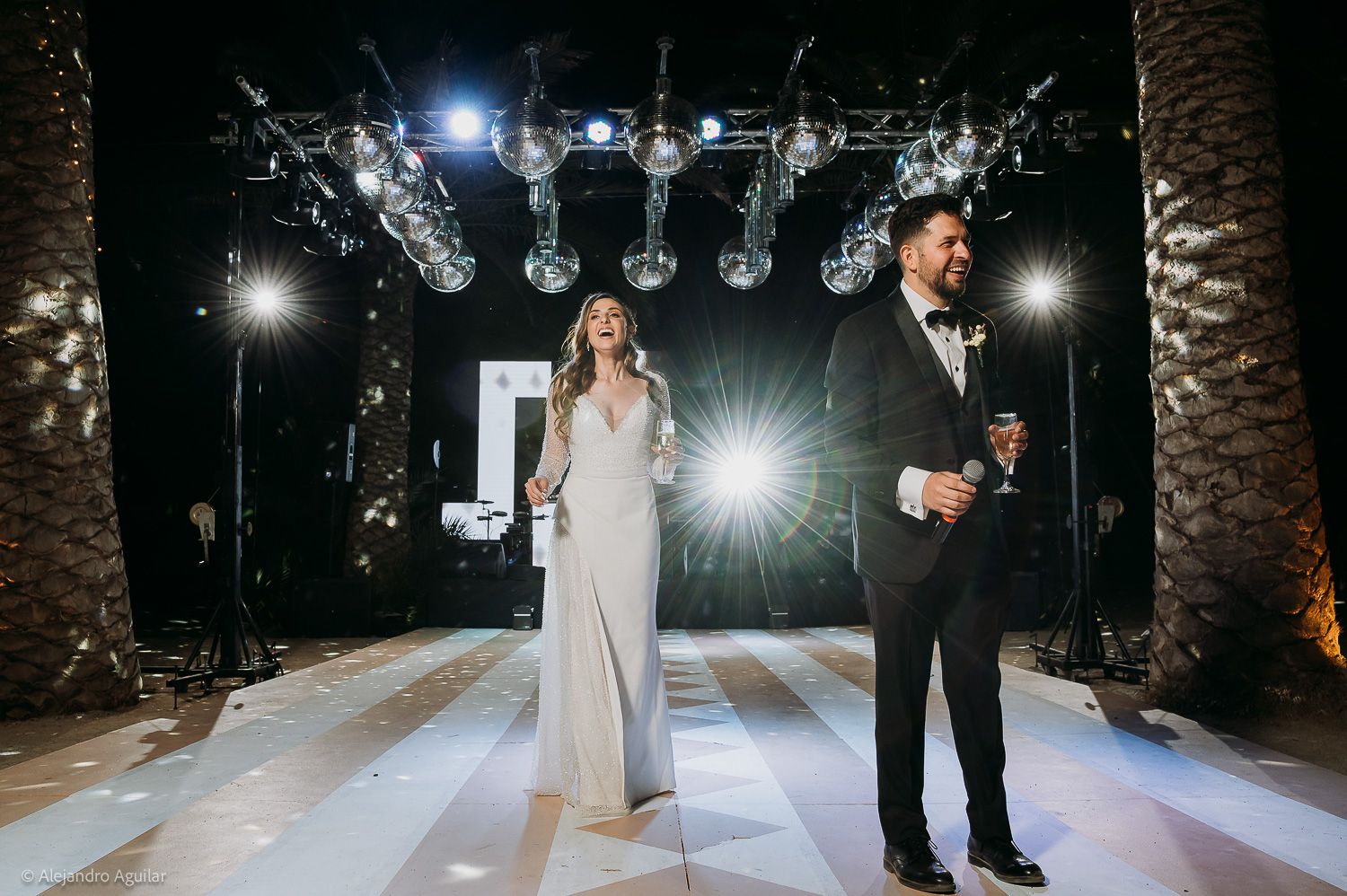 Elegant couple in formal attire standing on a dance floor at a wedding reception with dramatic lighting.