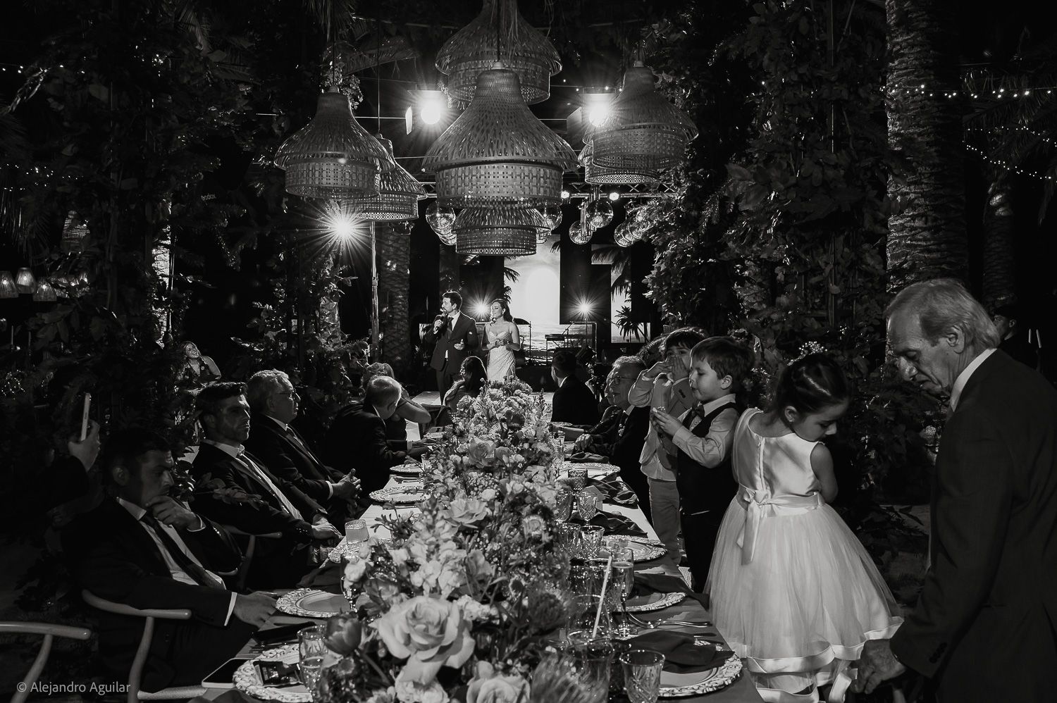 Black and white photo of elegant outdoor wedding reception dinner with guests seated at long decorated table under hanging lights.