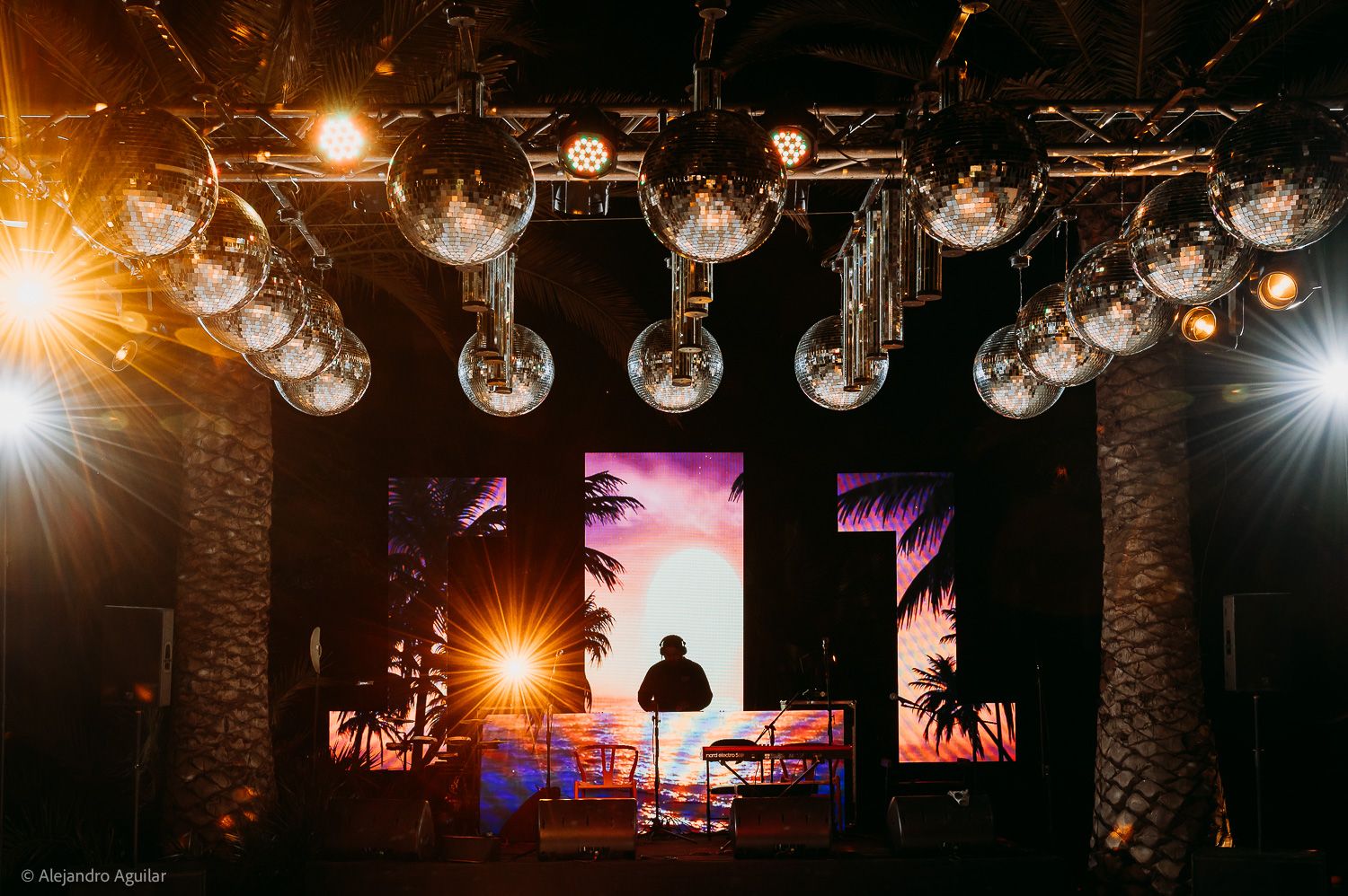 DJ performing on stage under globe lights with vibrant tropical sunset backdrop at an outdoor night event.