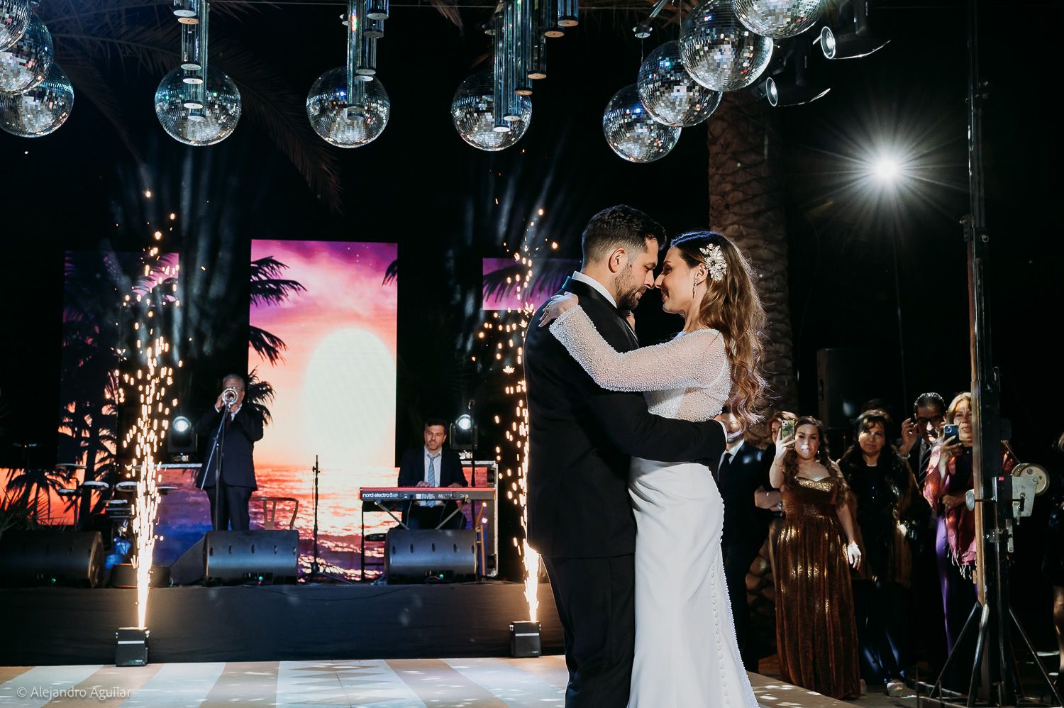 Bride and groom share first dance under disco balls with sparklers and guests at elegant wedding reception.