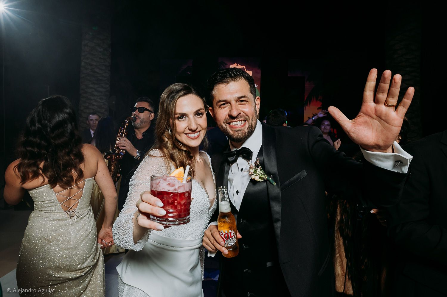Bride and groom smiling at wedding reception, holding drinks while celebrating on dance floor with guests in background.