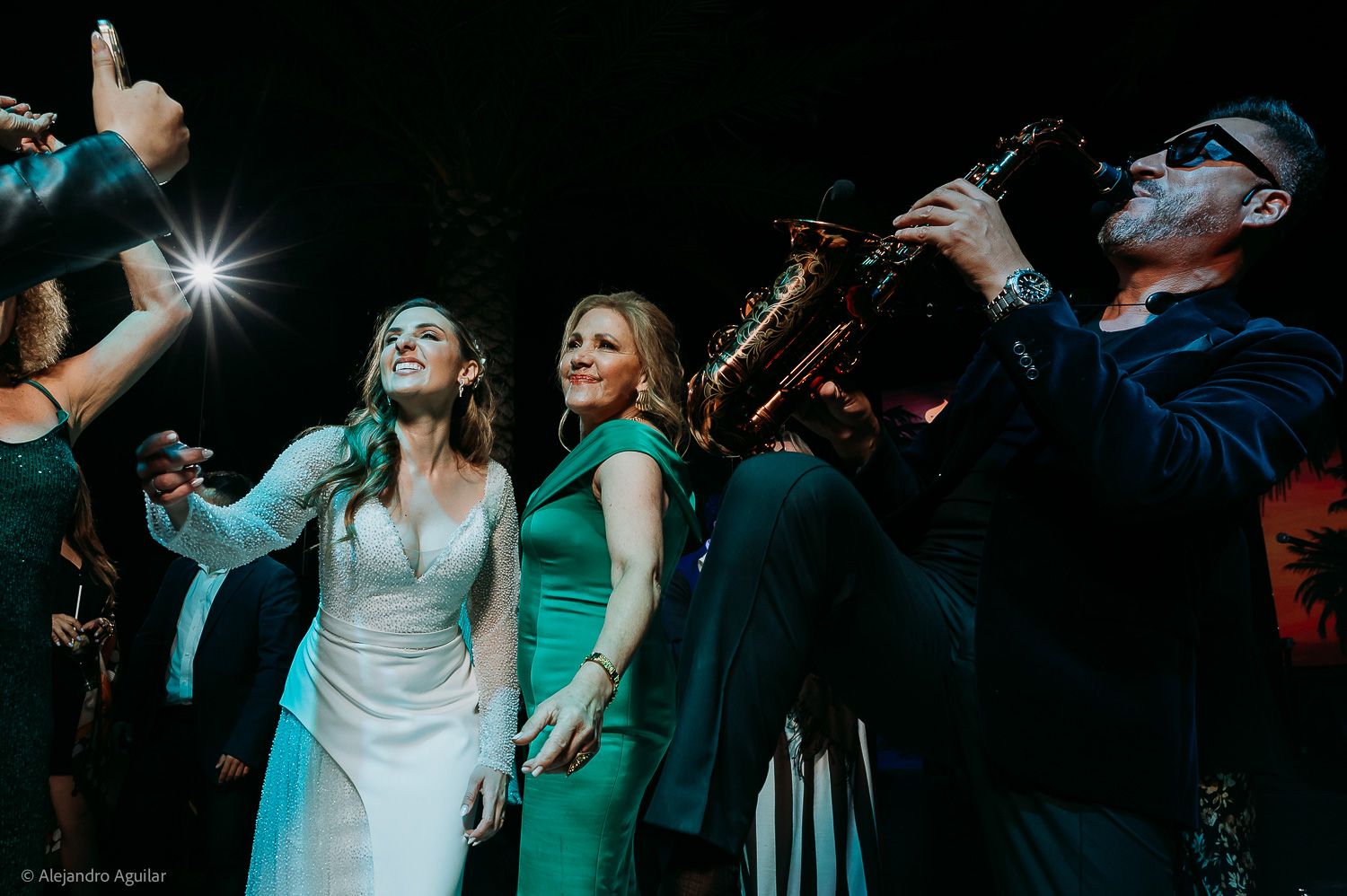 Bride and guests celebrate joyfully at wedding reception with live saxophone performance under dramatic stage lighting.