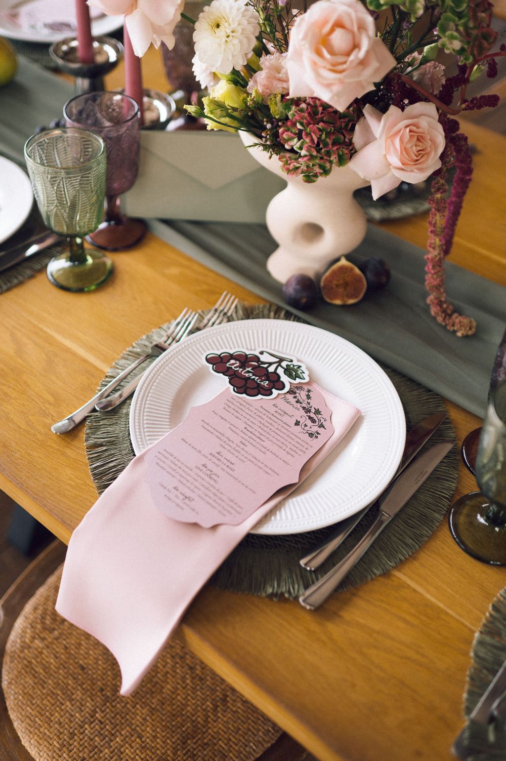 Elegant wedding table setting with pink napkin, floral menu card, white plate, and blush rose centerpiece in white vase.