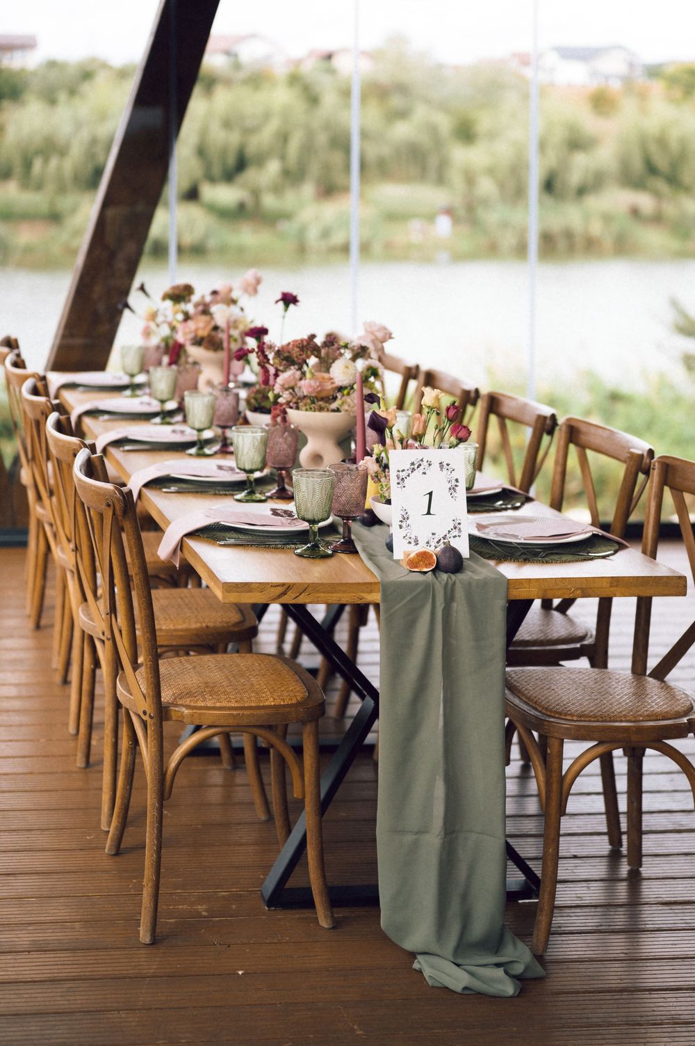 Elegant outdoor wedding reception with wooden cross-back chairs, sage green table runner, floral centerpieces, and table number cards.