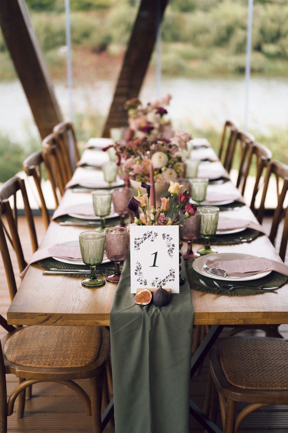 Elegant wedding table setting with sage green runner, dusty rose plates, green goblets, and floral centerpieces on a rustic wood table.