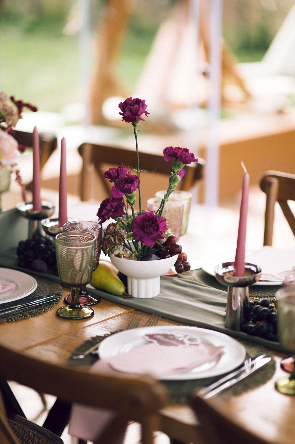 Elegant wedding table setting with purple carnations, pink taper candles, blush plates, and a green table runner on a wooden table.