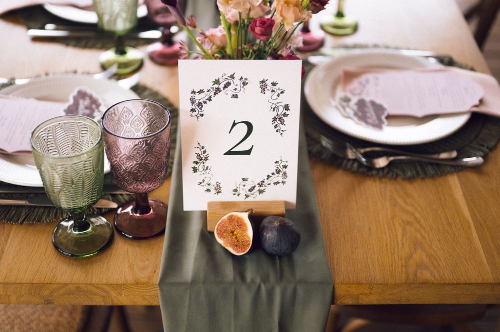 Elegant wedding table setting with table number 2 card, colorful glassware, floral centerpiece, and figs on a gray runner.