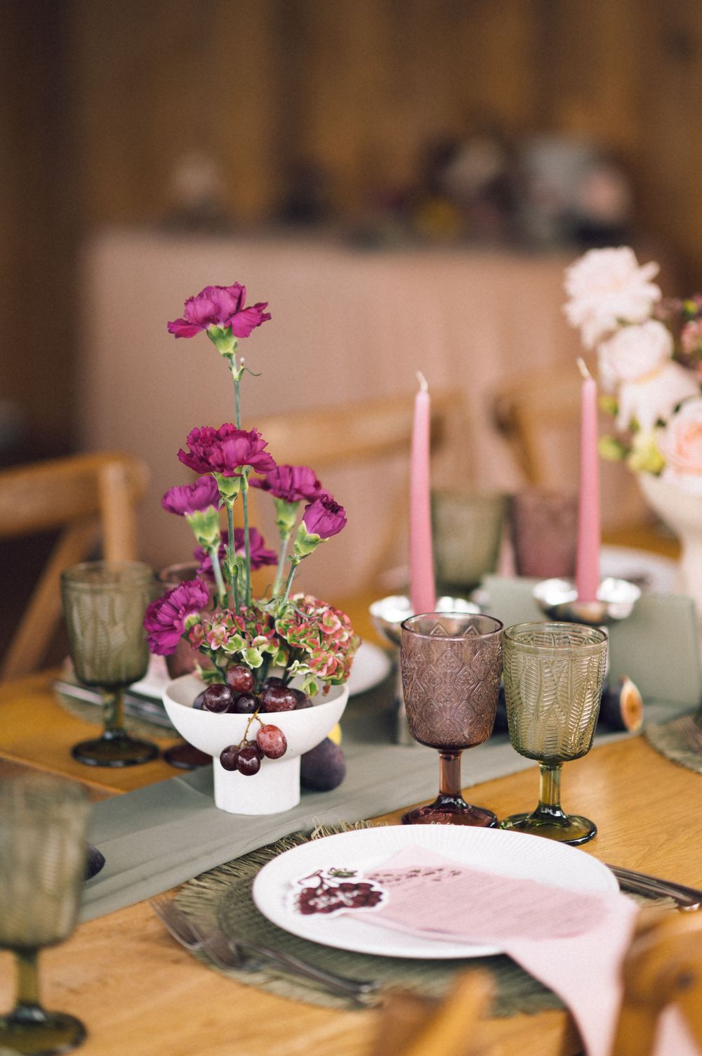 Elegant table setting with purple carnations, pink candles, and colorful glassware on a wooden dining table.