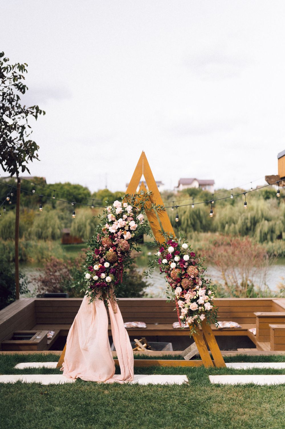 Elegant outdoor wedding arch with gold triangle frame, pink draping, and lush floral arrangements beside a serene pond.