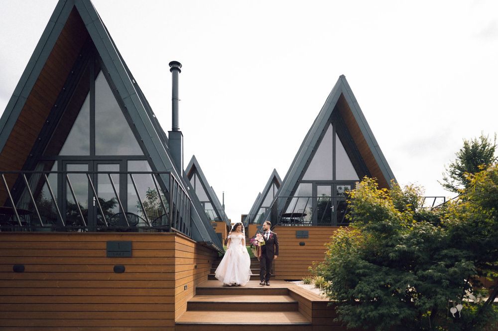 Bride and groom stand on steps outside A-frame glass wedding venue surrounded by lush greenery.