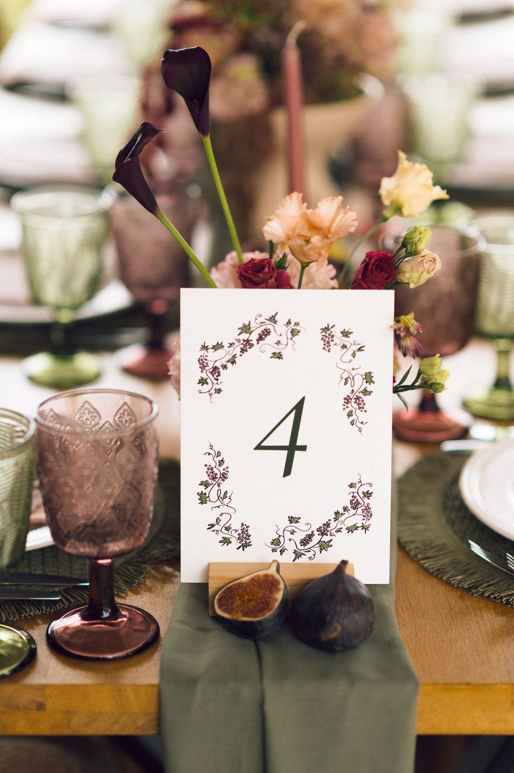 Elegant wedding table number 4 card with floral wreath, purple calla lilies, roses, green goblets, and figs on rustic table.