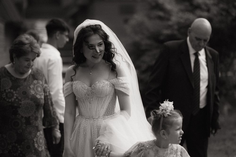 Bride in elegant tulle wedding gown and veil walks with flower girl and guests in black and white photo.