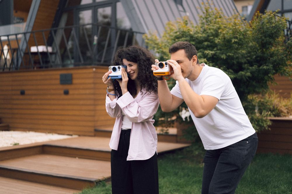 Two people smiling and taking photos with Polaroid cameras outdoors near a modern wooden building.