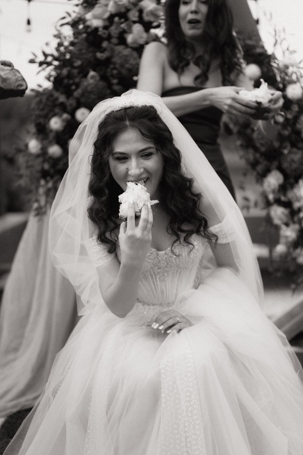 Black and white photo of a smiling bride in a veil and gown eating wedding cake at her reception.