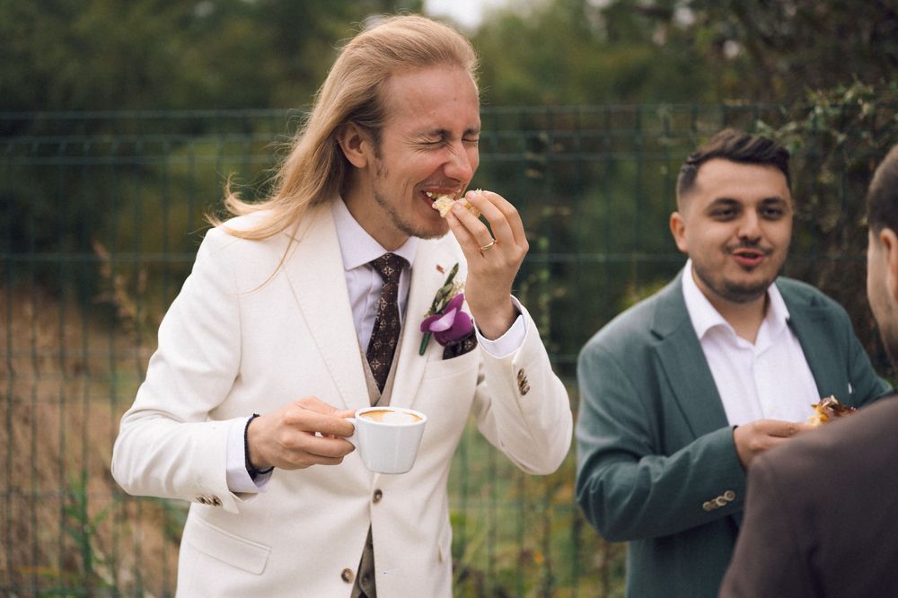 Man in white suit holding coffee cup and eating outdoors at a social gathering with guests in background.