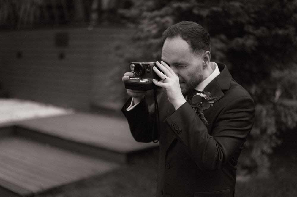 Black and white photo of a man in a suit photographing with a vintage camera outdoors.