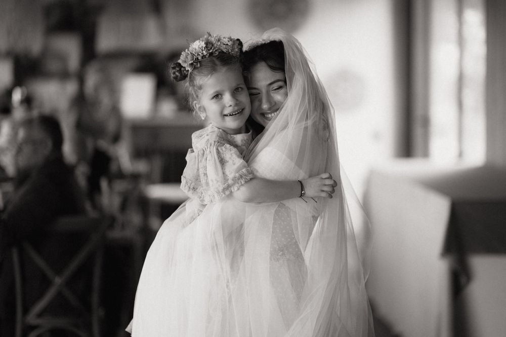 Bride in white veil hugs smiling flower girl in tulle dress, black and white wedding photo.