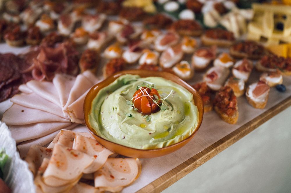 A buffet spread featuring a bowl of creamy green dip garnished with tomato, surrounded by assorted meats and appetizers.
