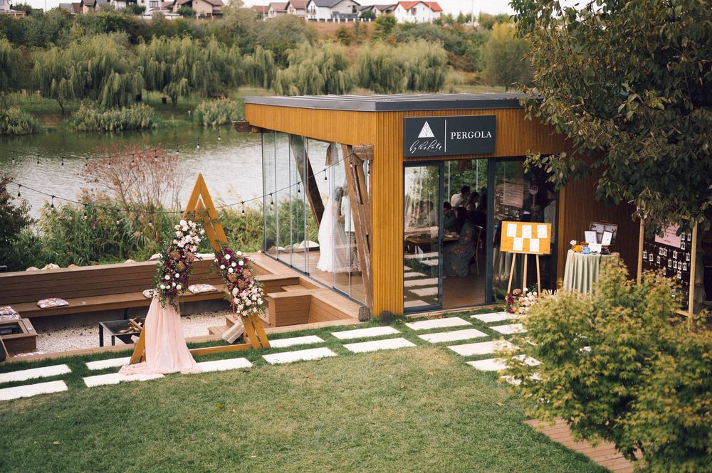 Modern pergola showroom with wooden exterior beside a lake, featuring floral arch decor and stepping stone pathway in a garden.