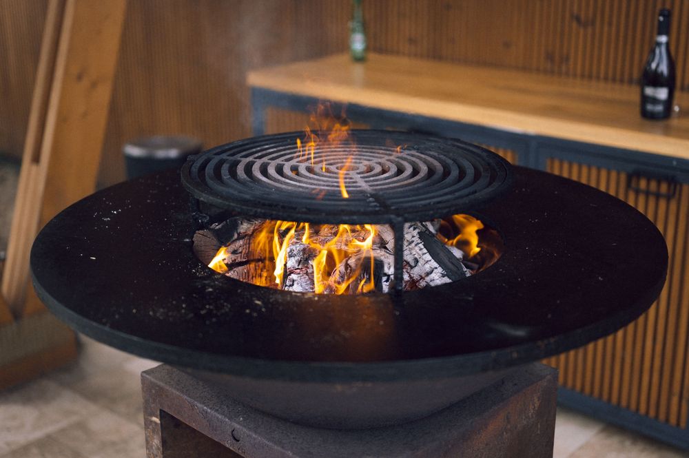 Outdoor fire pit grill with open flames and circular grate, set on a pedestal near wooden cabinetry.