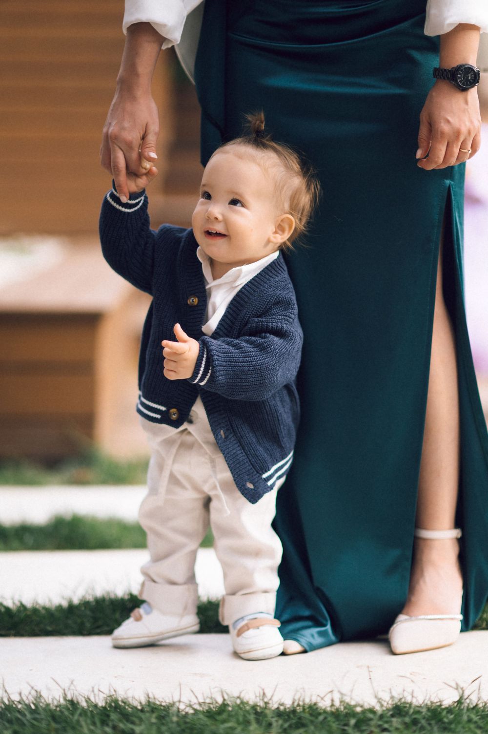 Adorable toddler in navy cardigan holds adult's hand while learning to walk outdoors near green dress.