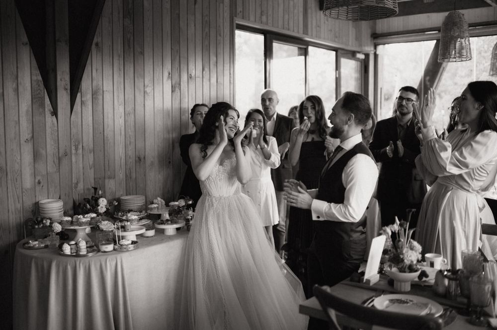 Black and white wedding reception photo of bride and groom sharing a joyful moment with guests clapping in rustic venue.