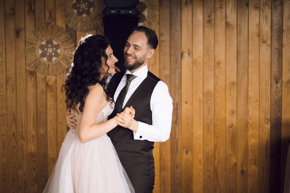 Bride and groom share first dance in elegant wedding attire, smiling at each other in front of wooden wall.