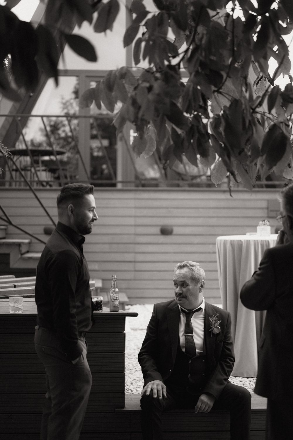 Black and white photo of two men in formal attire conversing outdoors at a wedding reception under leafy trees.