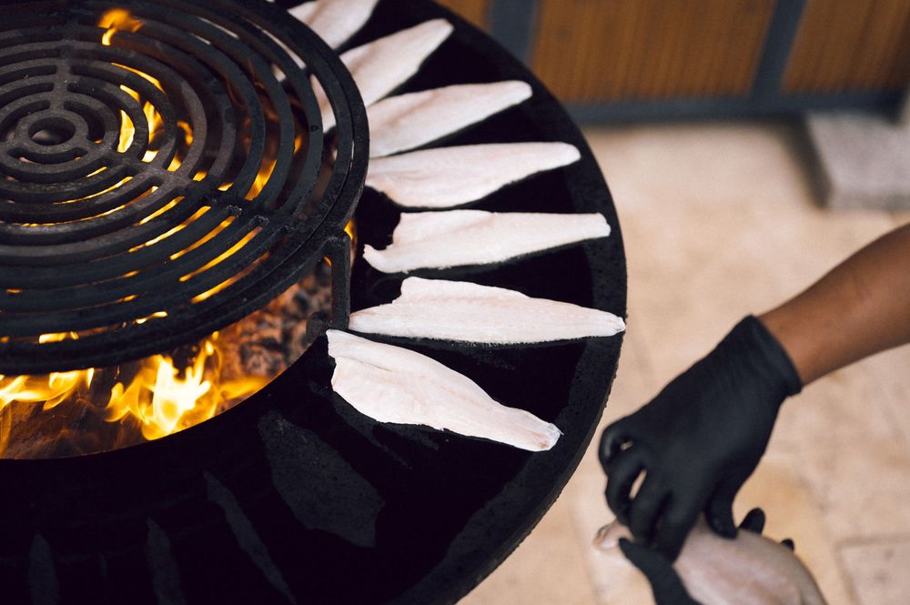 Chef grilling fresh white fish fillets on a round open-flame grill, wearing black gloves in a restaurant kitchen.