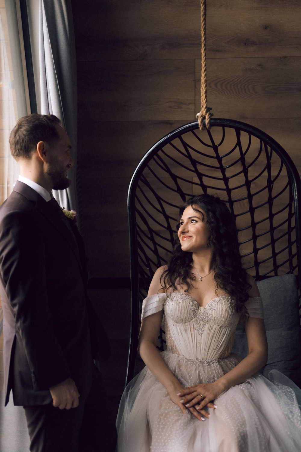 Bride in a white gown sits in a hanging wicker chair, smiling at a groom in a dark suit indoors.