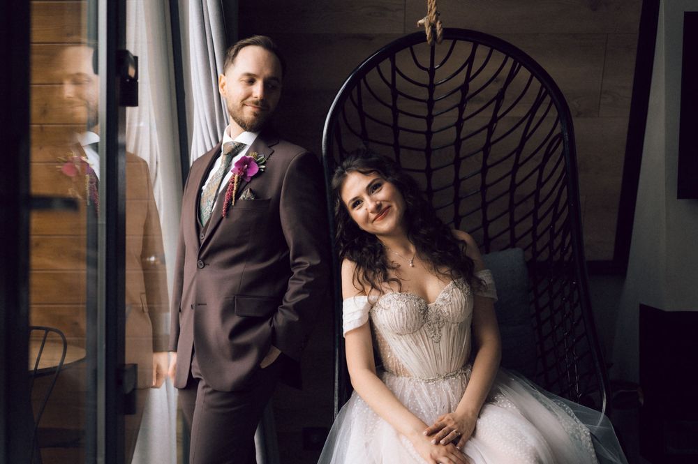 Bride in white gown seated in hanging chair beside groom in brown suit near window at wedding venue.