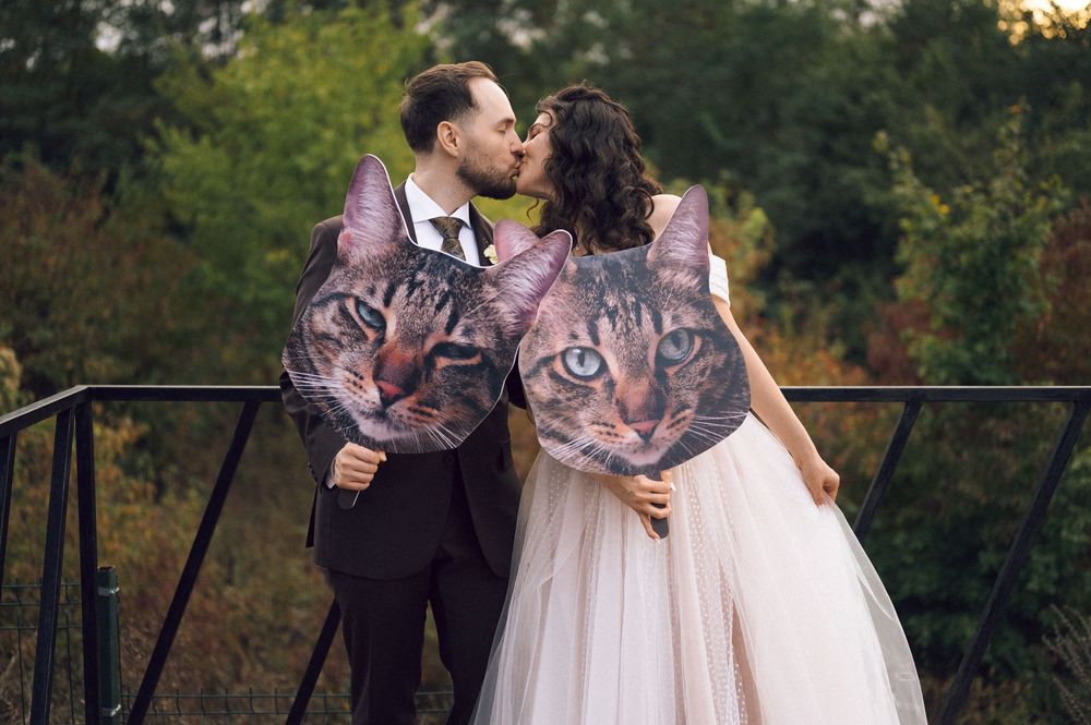 Wedding couple kissing while holding large cat face props outdoors, bride in white gown and groom in black suit.