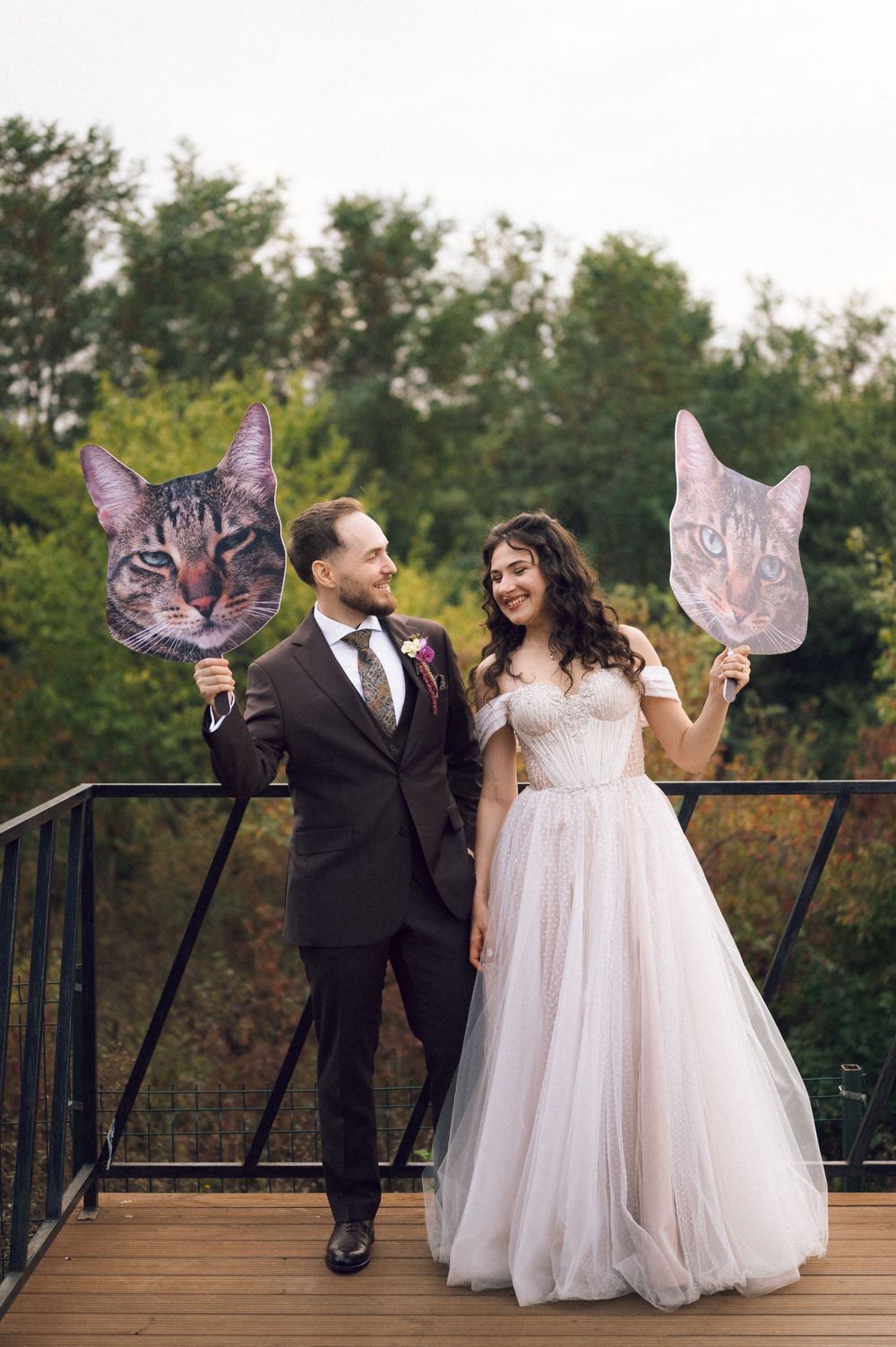 Bride and groom holding large cat face cutouts on a wooden deck, smiling at each other in wedding attire.