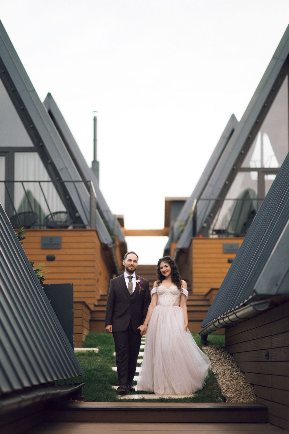 Bride and groom hold hands between modern A-frame cabins, she in a blush gown, he in a dark suit.