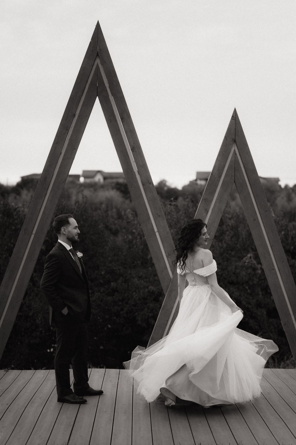Black and white wedding photo of bride in flowing gown and groom standing before geometric triangle sculptures on a deck.