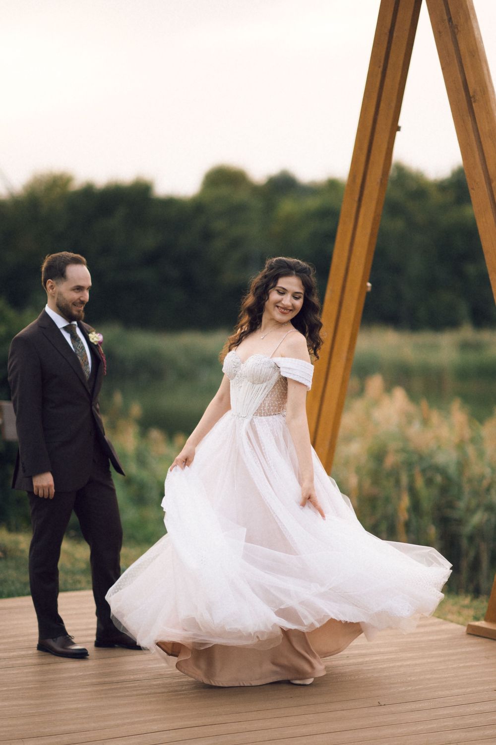Bride in flowing off-shoulder white gown twirls on wooden deck as groom watches during outdoor wedding celebration.
