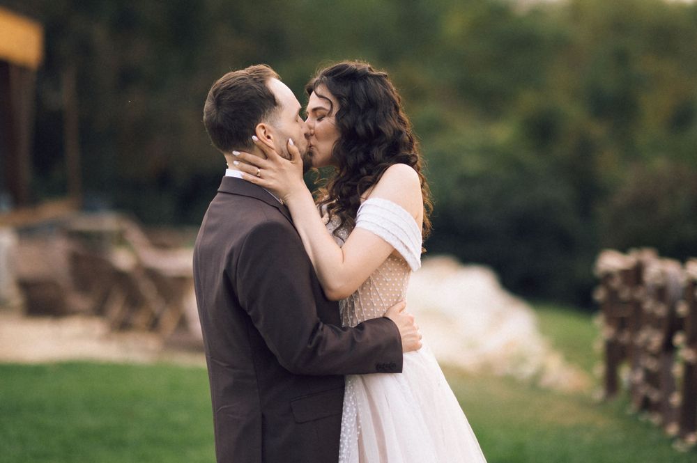 Couple sharing a romantic kiss outdoors in wedding attire, bride in white off-shoulder gown, groom in brown suit.