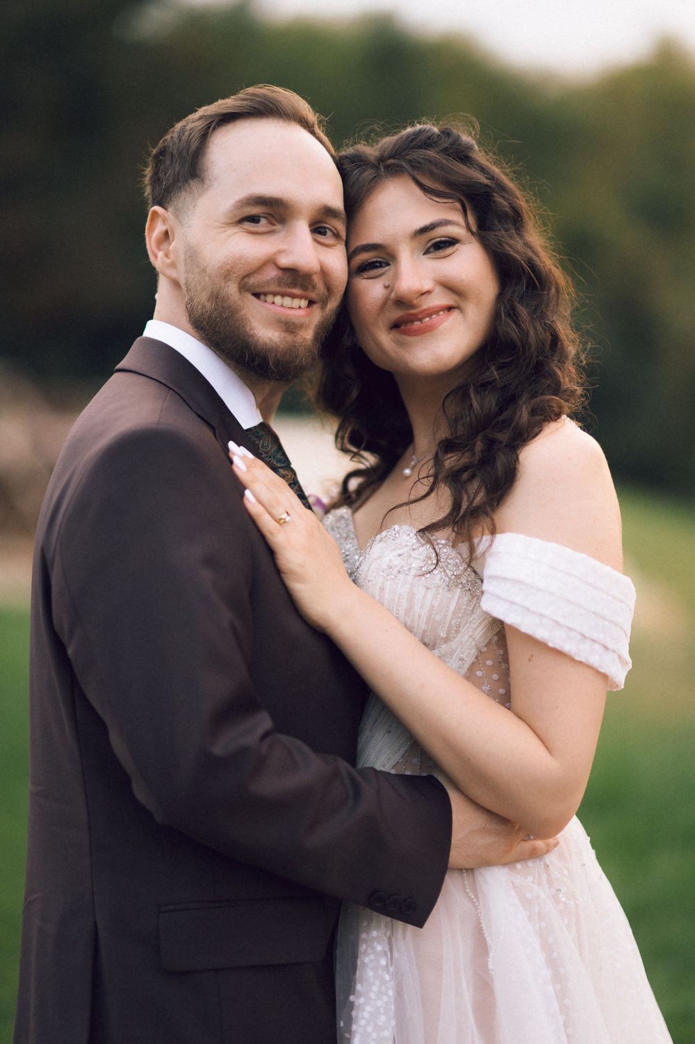 A smiling couple in wedding attire embraces outdoors, the bride in an off-shoulder gown and groom in a dark suit.