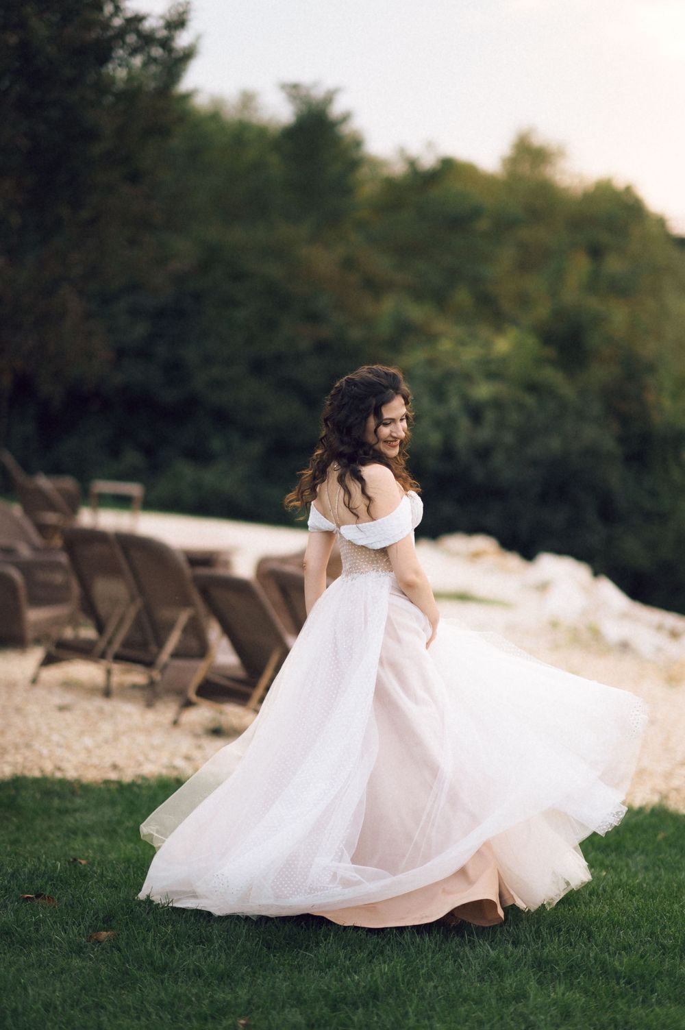 Smiling bride in off-shoulder white tulle gown twirling outdoors on green lawn with trees and chairs in background.