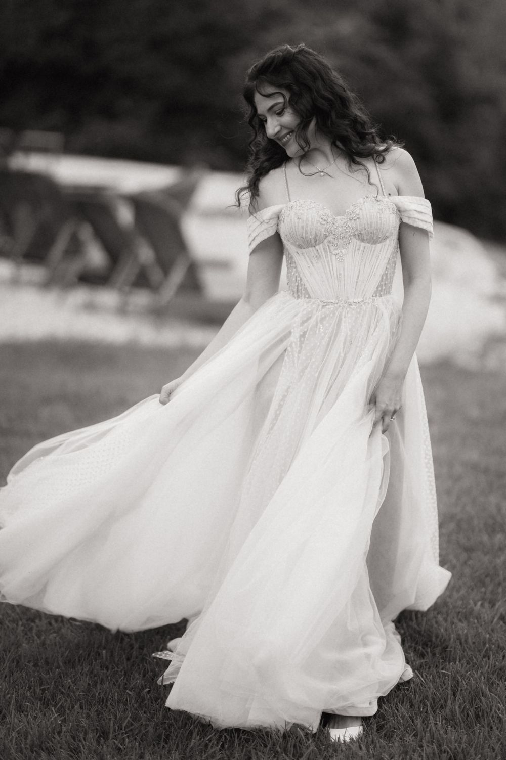 Bride in off-shoulder beaded wedding gown twirling on grass in black and white outdoor photo.