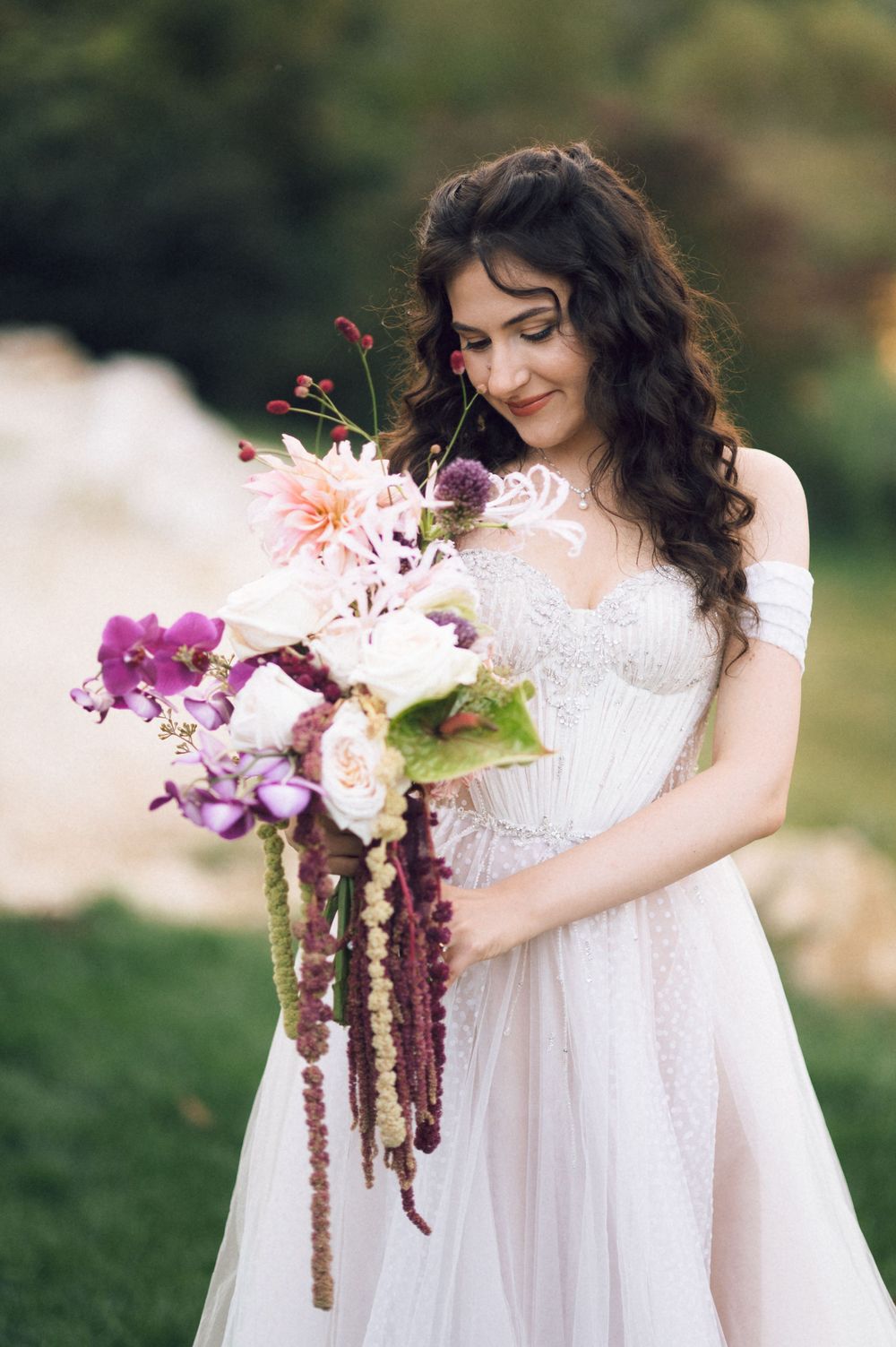 Bride in white lace gown holding a lush bouquet of purple orchids, pink dahlias, and trailing amaranthus outdoors.