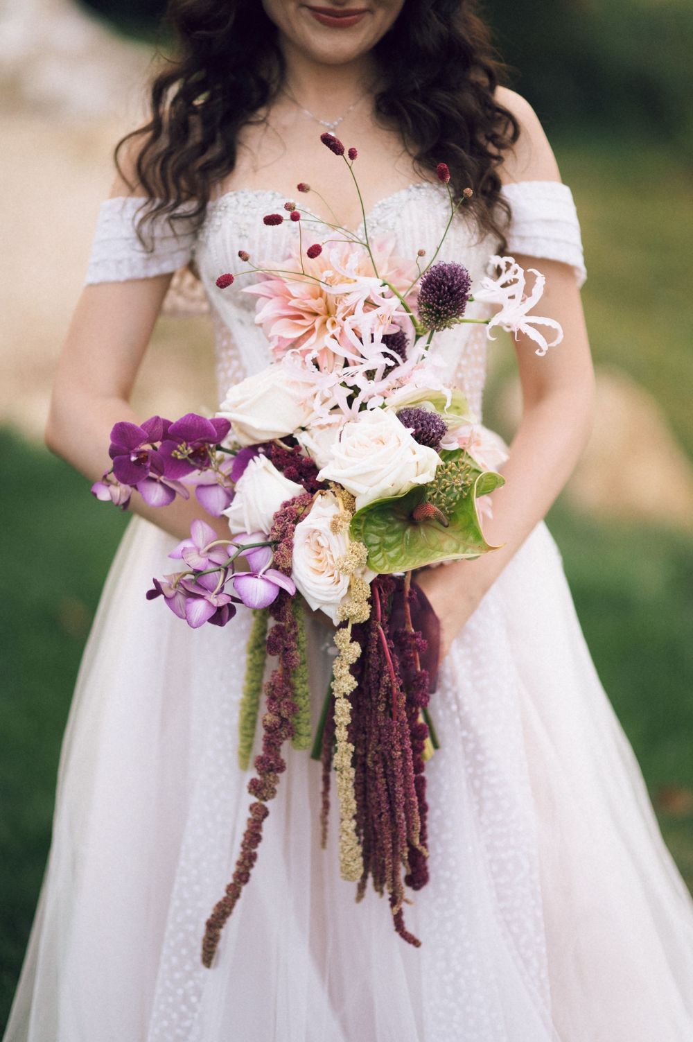 Bride in off-shoulder white gown holds cascading bouquet with purple orchids, roses, thistles, and amaranthus.