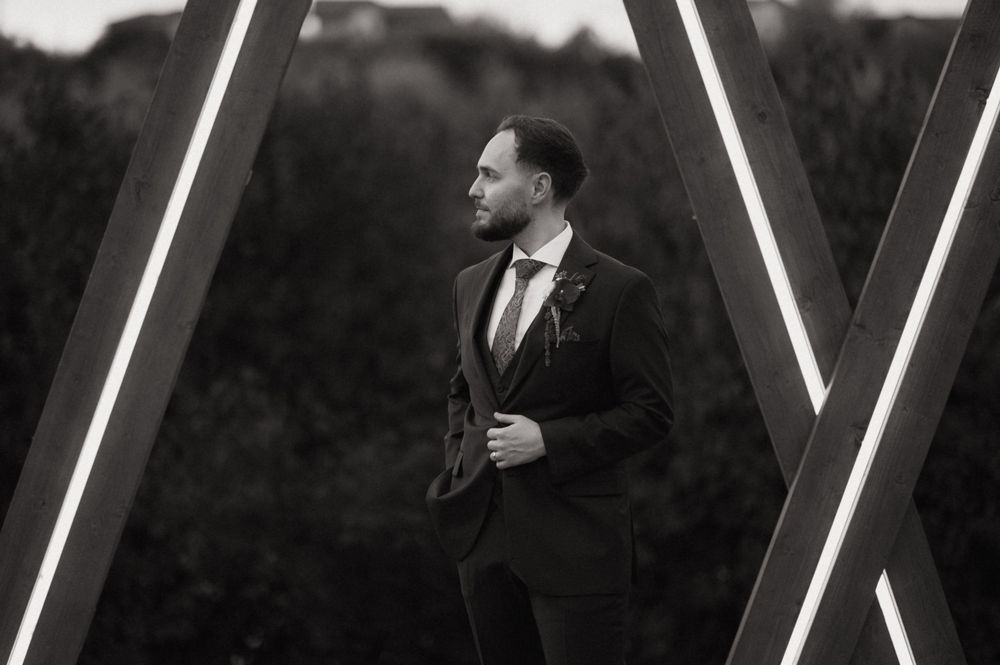 Elegant man in dark suit poses between geometric wooden frames in black and white portrait photography.