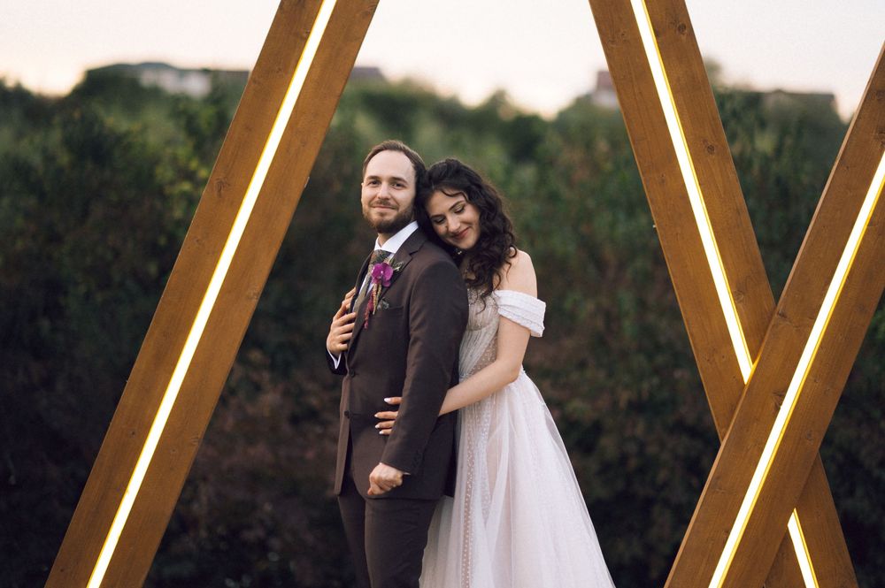 Bride and groom posing between illuminated wooden arch frames with lush green trees in the background.