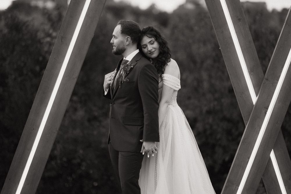 Black and white wedding photo of bride and groom standing back-to-back beneath geometric wooden arch with LED lights.