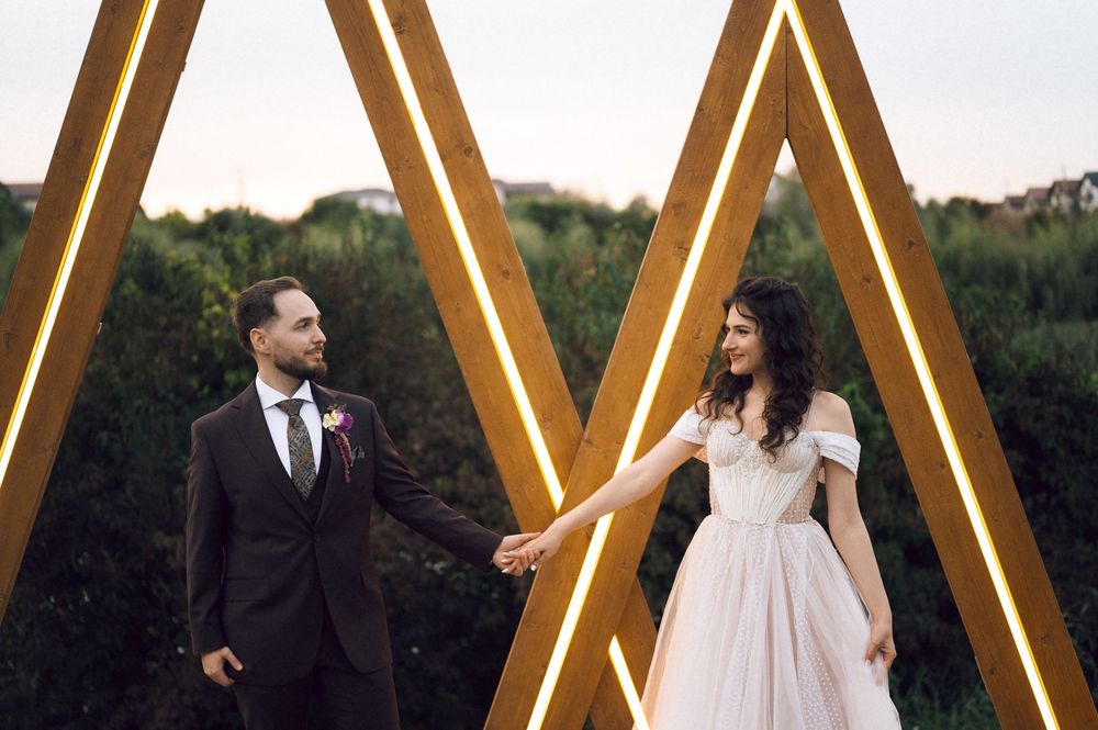 Bride and groom hold hands in front of illuminated wooden triangle arch outdoors on their wedding day.