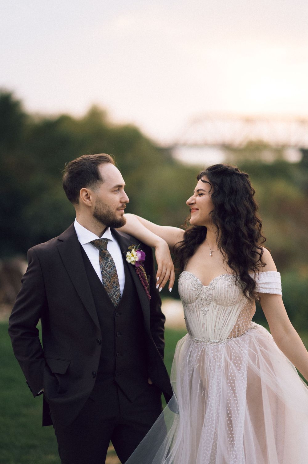 A bride and groom share a romantic moment outdoors at sunset, with the bride in a white gown resting her arm on the groom.