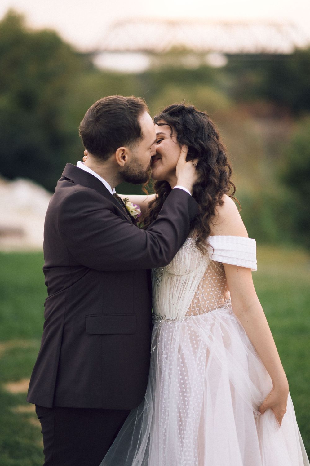 A groom in a dark suit tenderly kisses his bride in an off-shoulder white gown outdoors near a bridge.