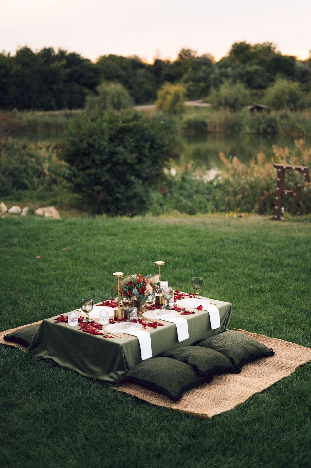 Romantic outdoor low table setting on green grass with dark cushions, red rose petals, candles, and a lake backdrop.
